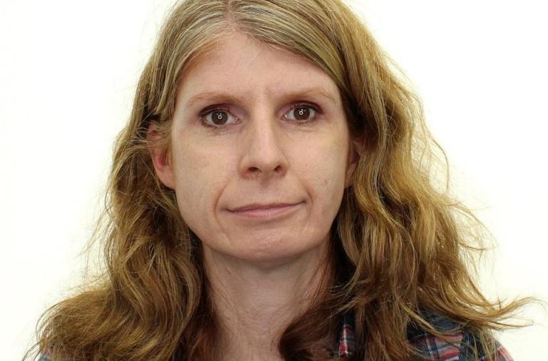 a headshot of Louise Simpson who has long wavy hair and brown eyes, she is wearing a blue and red check shirt